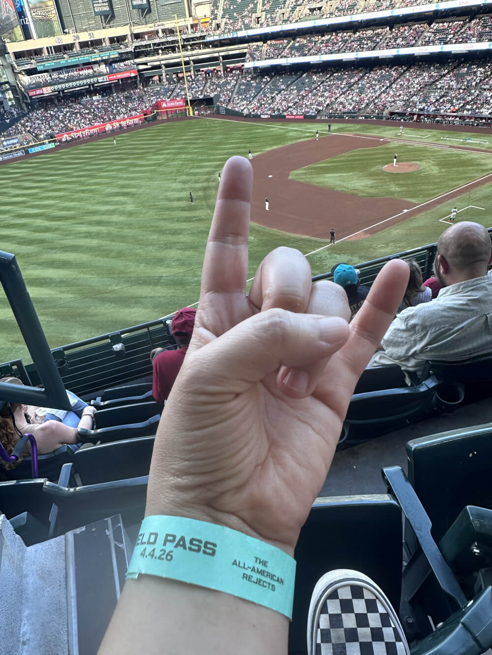 Hand giving devil horns wearing a blue wristband Field Pass that says the date and The All-American Rejects. The Diamondbacks are playing on Chase Field in the background