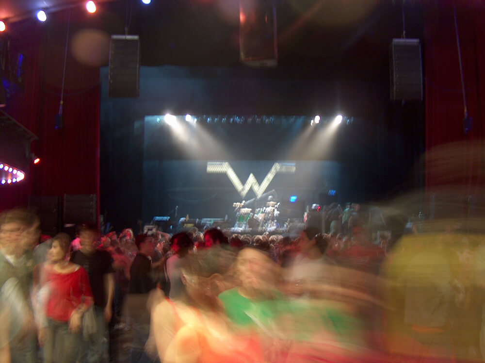 the Weezer W on the stage as blurry people move around the venue